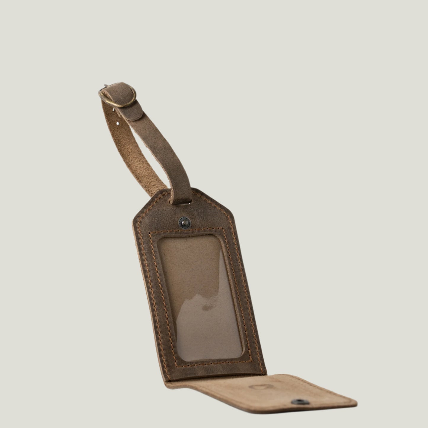 Close-up of a Sand-colored Bond Leather Luggage Tag with antique brass hardware, a clear ID sleeve, and a personalized fire-branded initial, set on a rustic surface.