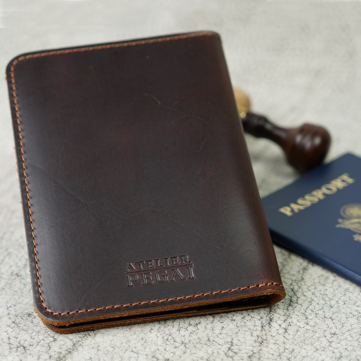 Close-up of the Pike Leather Passport Wallet in Djor Brown, resting atop a textured surface beside a U.S. passport and wax stamp. Embossed with the Atelier PEGAI logo, the full-grain leather reveals natural markings—celebrating the beauty of imperfection and timeless handmade design.