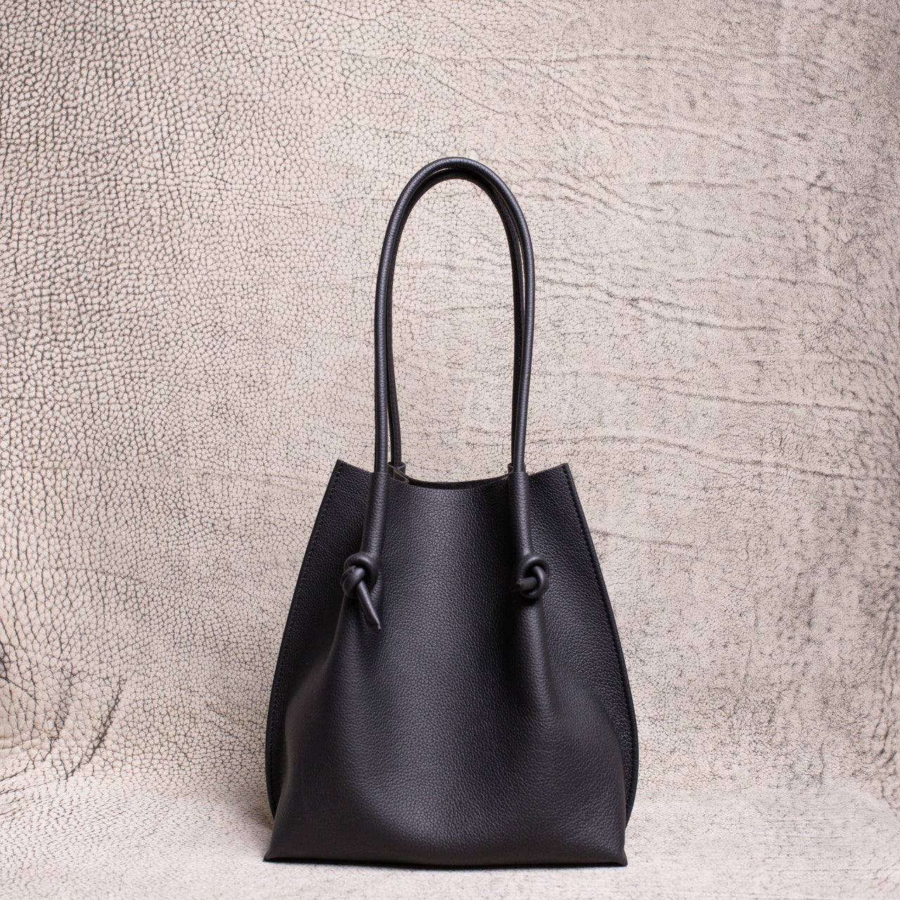 Front view of the Flora Everyday Tote in black Togo leather, showcasing its softly structured silhouette and knotted handle details against a textured neutral backdrop.