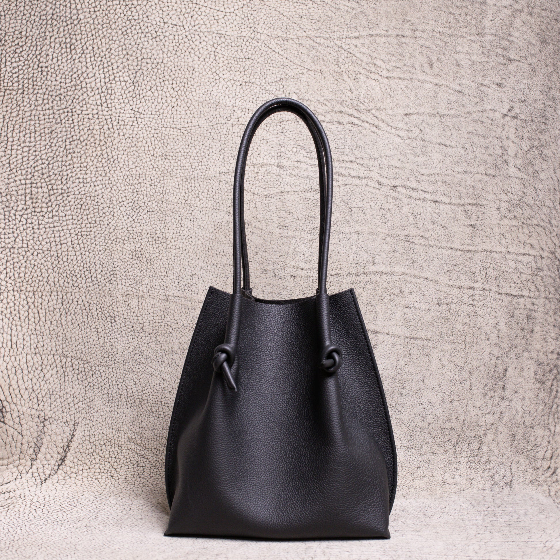 Front view of the Flora Everyday Tote in black Togo leather, showcasing its softly structured silhouette and knotted handle details against a textured neutral backdrop.