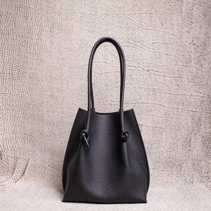 Front view of the Flora Everyday Tote in black Togo leather, showcasing its softly structured silhouette and knotted handle details against a textured neutral backdrop.