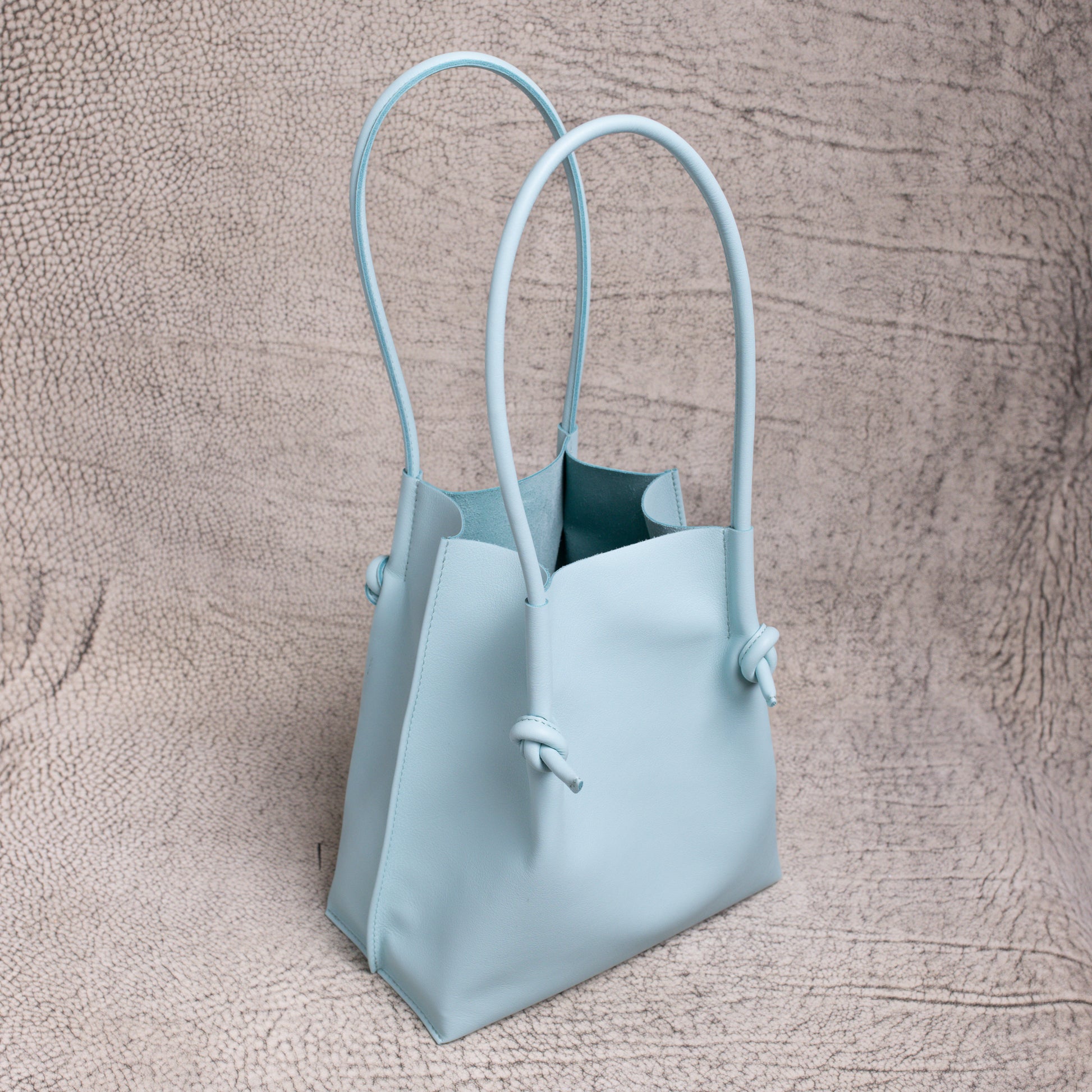 A side-angle view of the Flora Everyday Tote in Silky Calf Sky, showing its sculptural shape and long rolled leather handles.