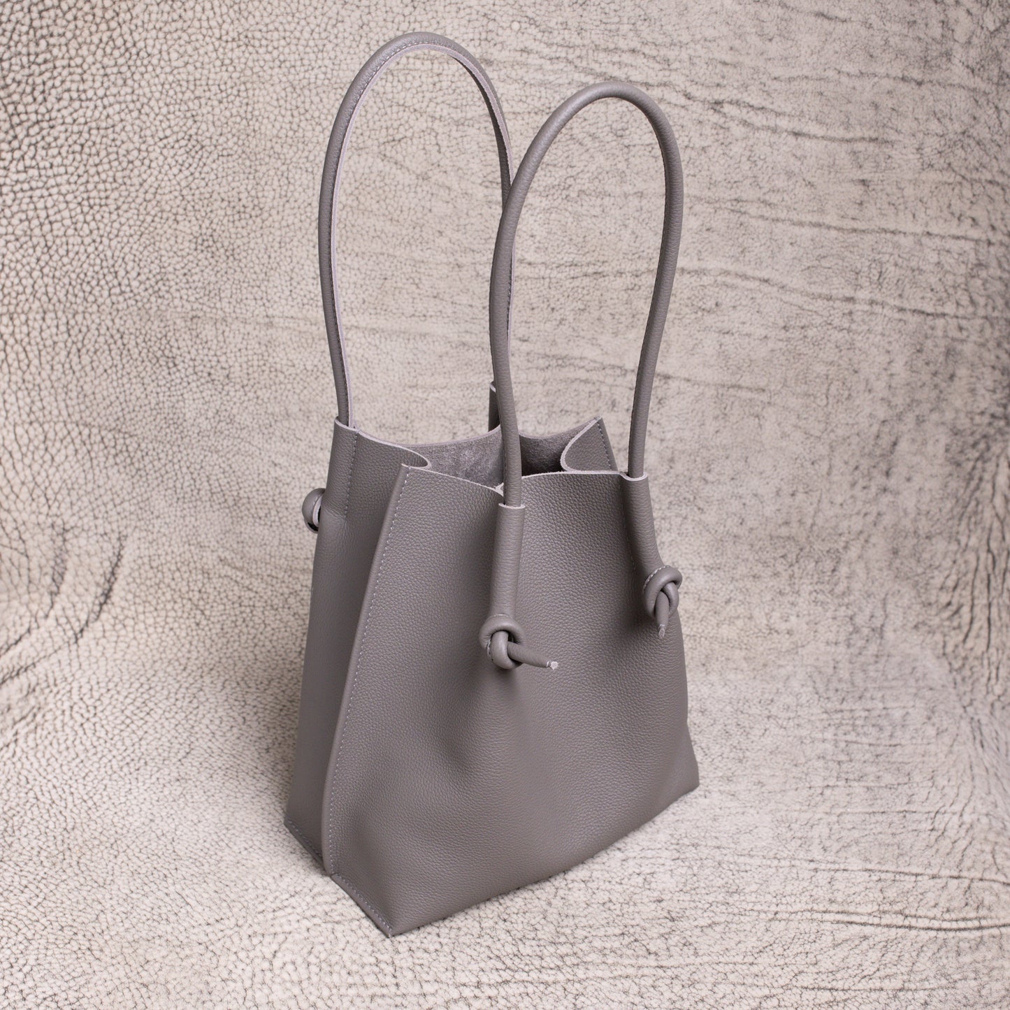 Three-quarter angle of the gray Flora tote, highlighting its sculpted silhouette and rolled handles against a neutral textured backdrop.