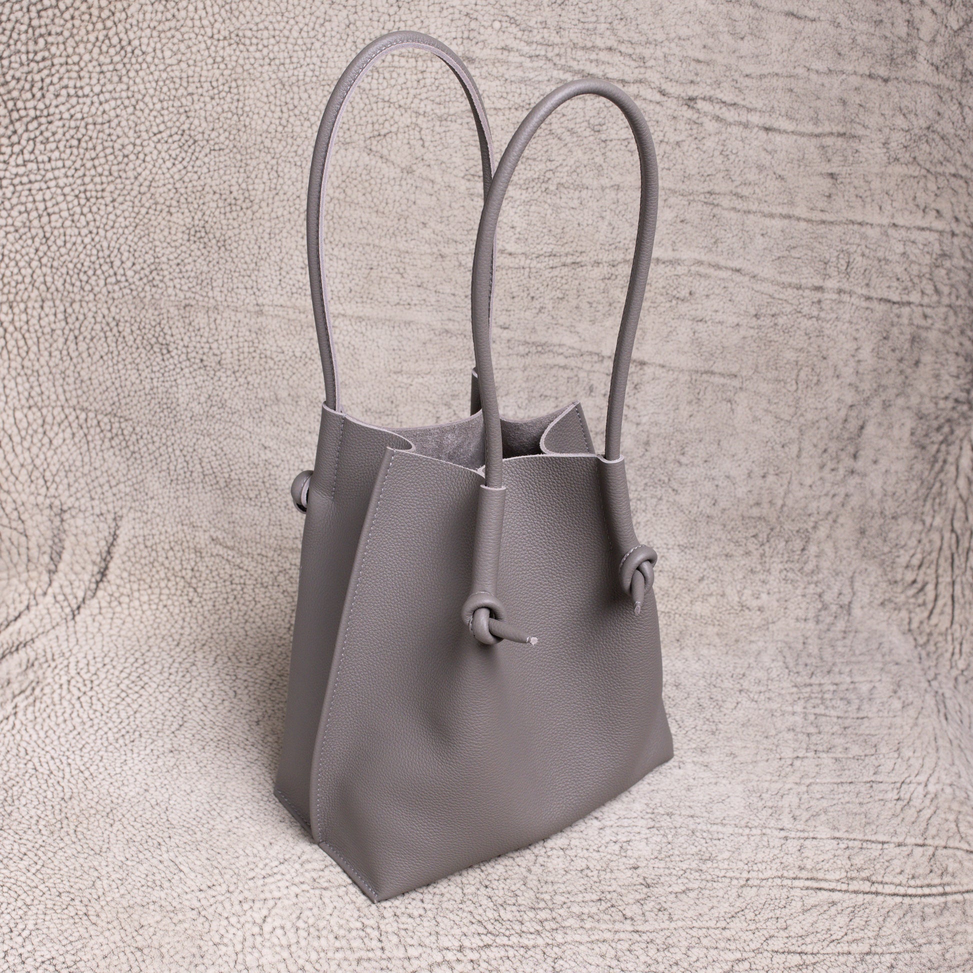 Three-quarter angle of the gray Flora tote, highlighting its sculpted silhouette and rolled handles against a neutral textured backdrop.