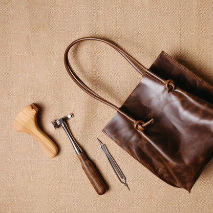 The Flora Everyday Tote laid flat with leatherworking tools beside it, reflecting Atelier PEGAI’s handcrafted roots and raw suede interior.