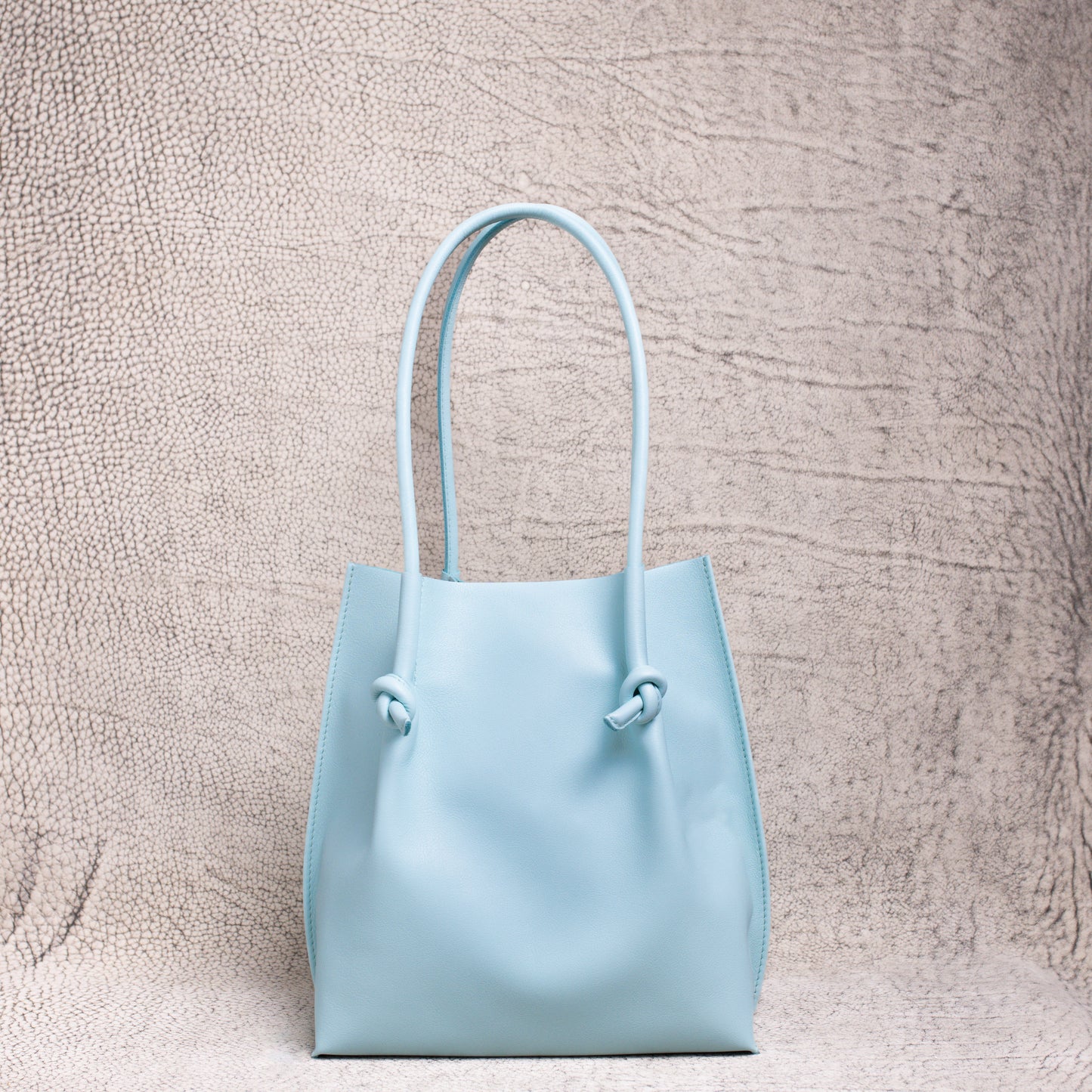 A minimalist light blue leather tote bag with knotted handle detailing, standing upright against a textured neutral backdrop.
