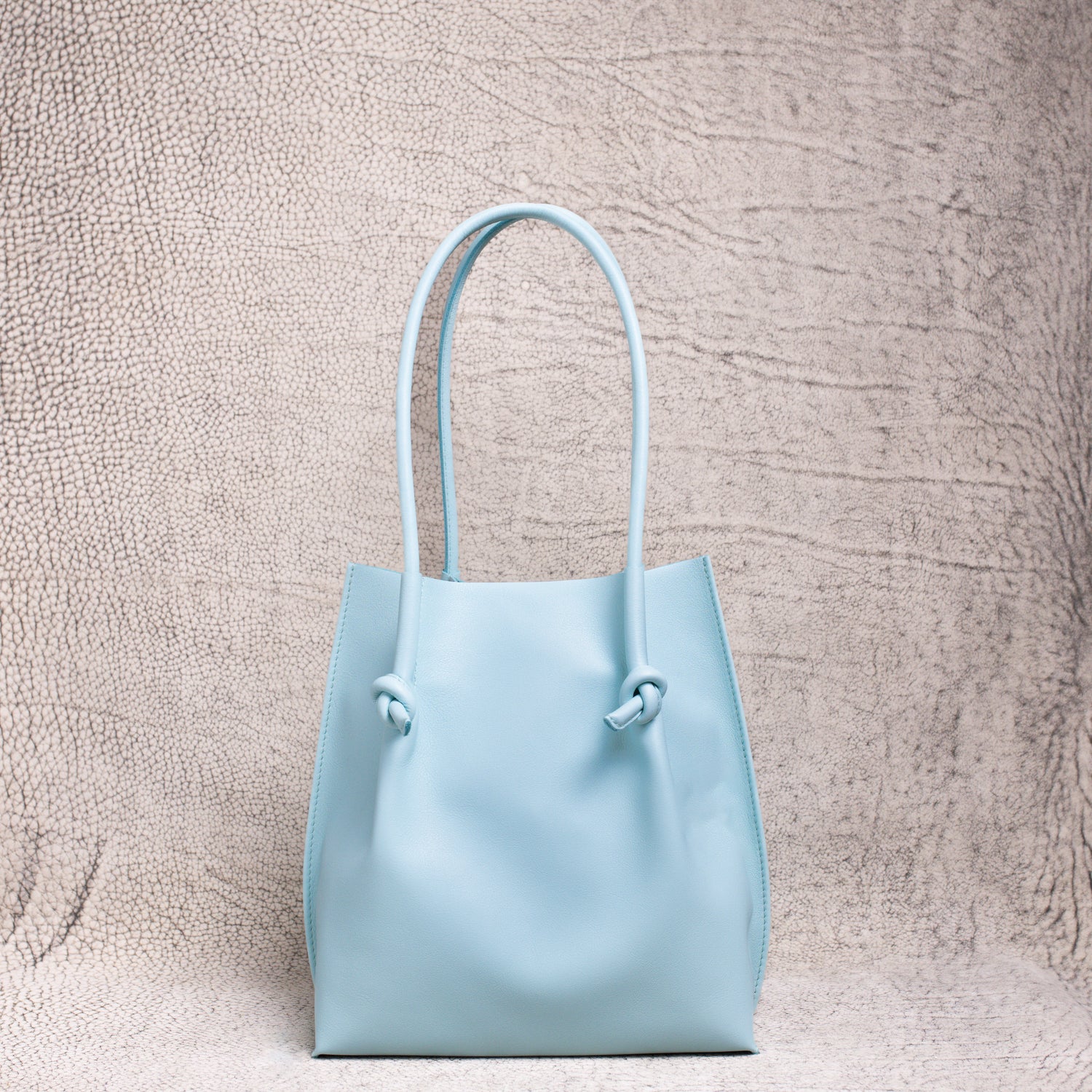 A minimalist light blue leather tote bag with knotted handle detailing, standing upright against a textured neutral backdrop.