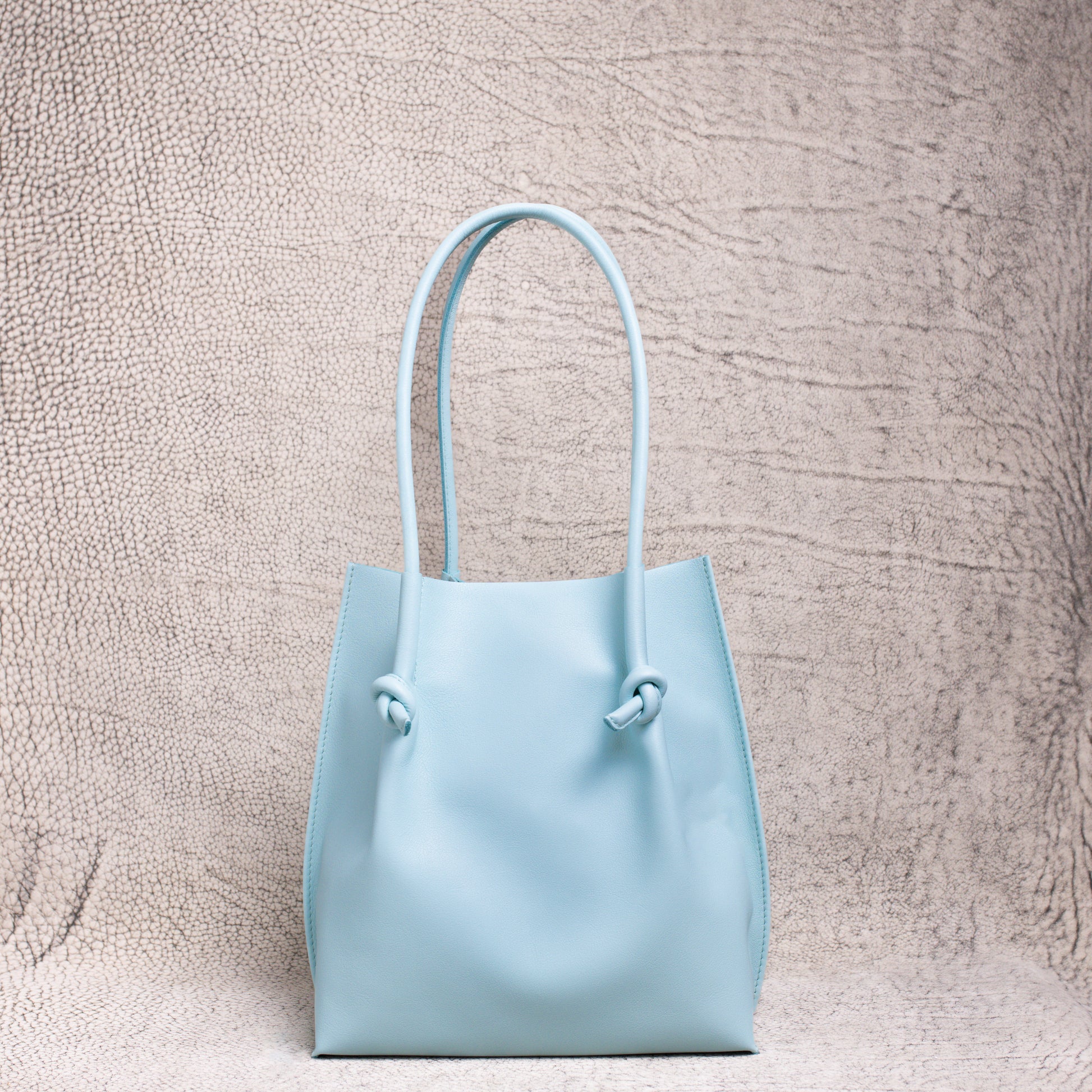 A minimalist light blue leather tote bag with knotted handle detailing, standing upright against a textured neutral backdrop.