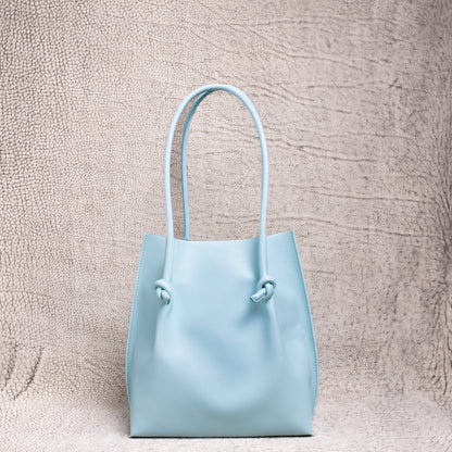 A minimalist light blue leather tote bag with knotted handle detailing, standing upright against a textured neutral backdrop.