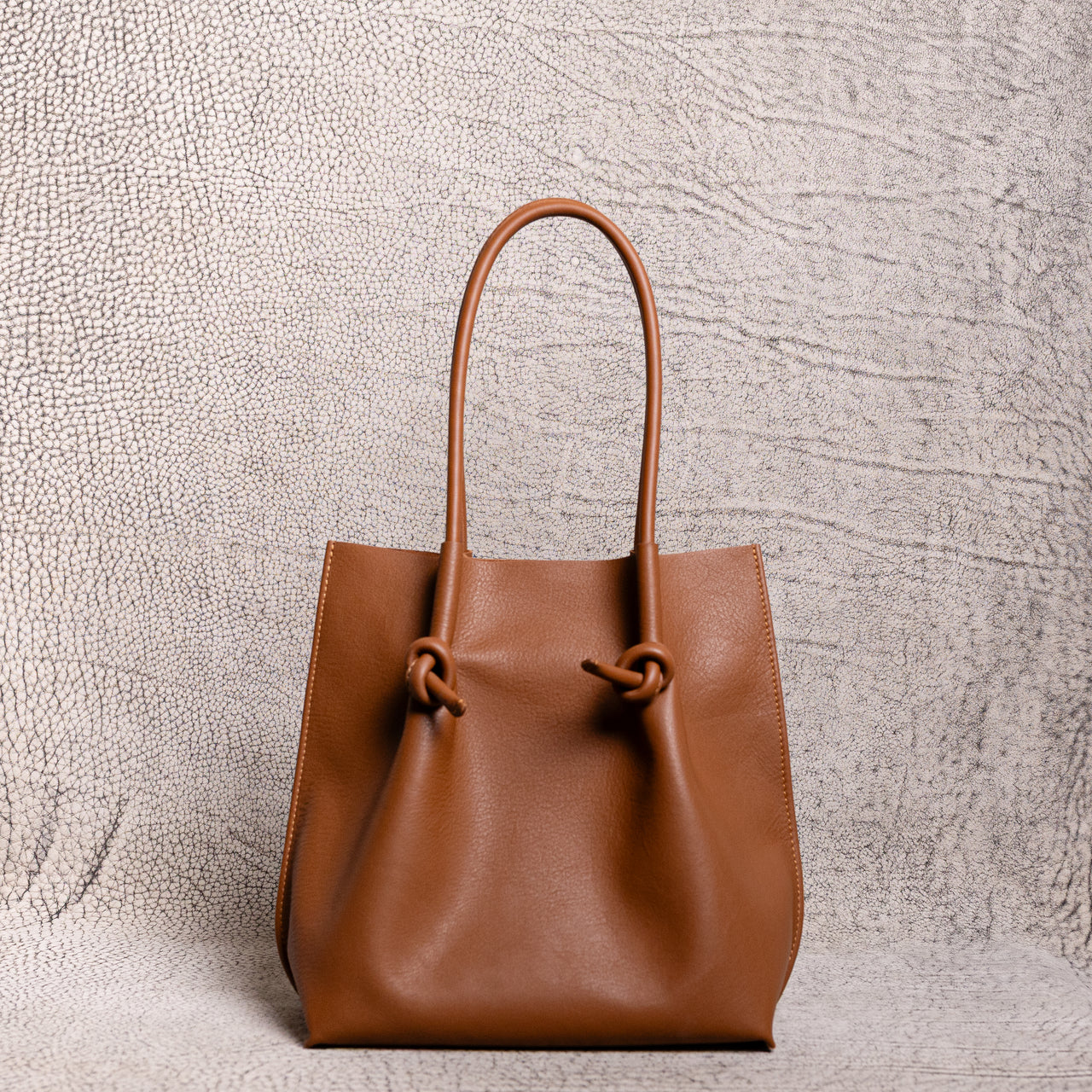 Front-facing view of the tan (taba) Flora tall leather tote bag on a textured background, highlighting its structured base, clean lines, and knotted handle details.
