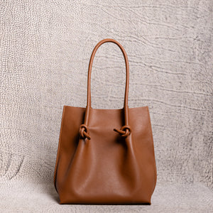 Front-facing view of the tan (taba) Flora tall leather tote bag on a textured background, highlighting its structured base, clean lines, and knotted handle details.