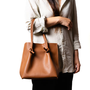 A woman wearing a cream blouse carries the tan (taba) Flora tall tote bag on her forearm, showcasing the bag’s compact vertical silhouette and double-handle design.