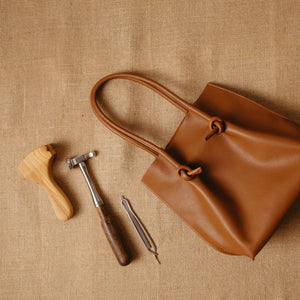 The tan (taba) Flora tall tote bag laid on a burlap surface next to traditional leathercraft tools, representing handmade quality and artisan craftsmanship.