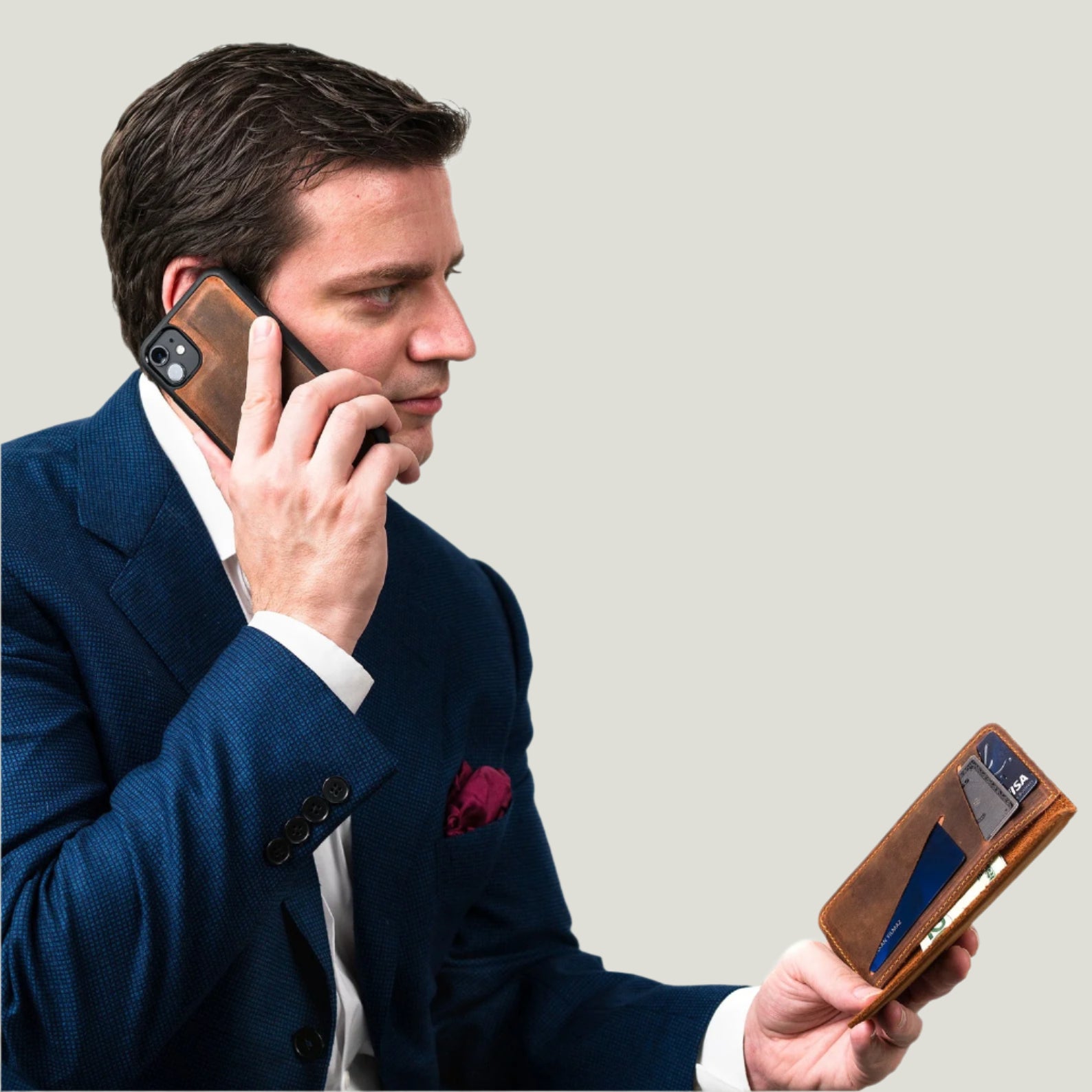 Man in navy suit using mahogany leather iPhone wallet case, holding detachable phone cover to ear while viewing card slots and cash compartment.